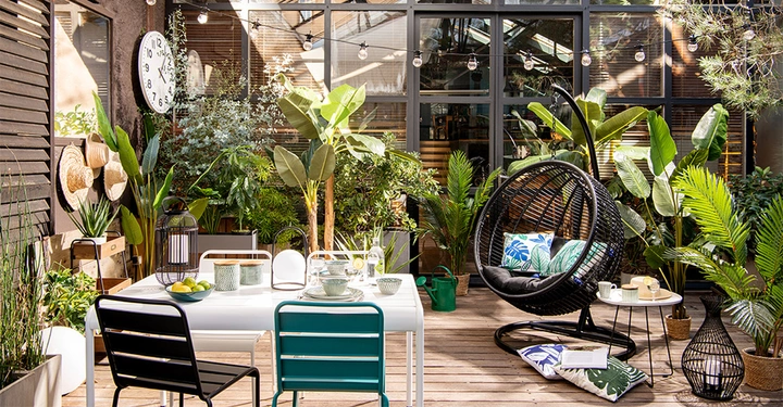 Galerie-Bild 5: Ein gedeckter Tisch mit Geschirr und Limetten steht auf einer Terrasse umgeben von vielen Pflanzen Dekorationen und einem runden Hängestuhl Lampenketten hängen oben eine große Uhr an der Wand.