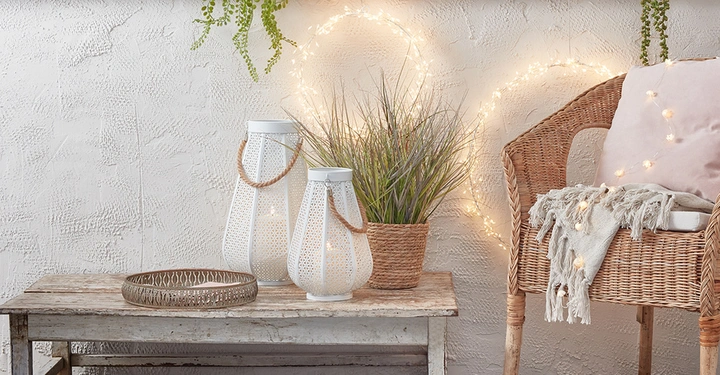Zwei weiße Laternen und ein Blumentopf stehen auf einem Holztisch in einem gemütlichen Raum Dekoration mit Lichterketten und einem Korbstuhl mit Kissen daneben.