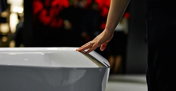 Hand berührt den RAND einer glänzenden weißen Wanne in einem AUSSTELLUNGSRAUM mit verschwommenen Menschen und roten Dekorationen im Hintergrund