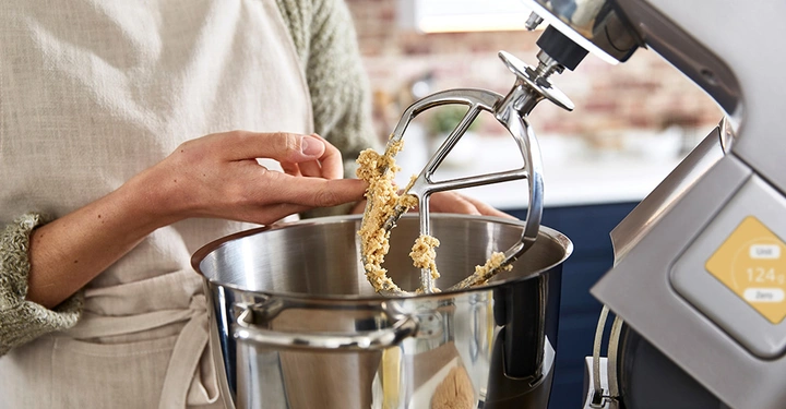 Frauenhände streifen Schokoteig von Rührstab einer geöffneten Kenwood Küchenmaschine