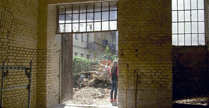 Galerie-Bild 2: Eine Person steht im Freien neben einem alten, baufälligen Gebäude aus Ziegelsteinen im Innenhof, in den Umbaumaterialien und Pflanzen zu sehen sind.