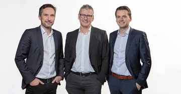 Drei Männer stehen vor einem weißen Hintergrund. Sie tragen Anzüge mit hellblauen Hemden und lächeln in die Kamera. Alle drei haben ihre Hände in den Hosentaschen.