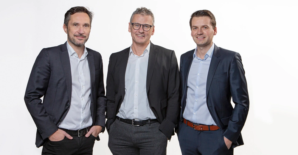 Drei Männer stehen vor einem weißen Hintergrund. Sie tragen Anzüge mit hellblauen Hemden und lächeln in die Kamera. Alle drei haben ihre Hände in den Hosentaschen.