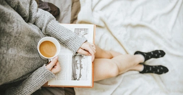 Person liest ein Buch und hält eine Tasse Kaffee. Sie trägt einen grauen Pullover und schwarze Socken. Sie sitzt auf einem hellen, weichen Teppich.
