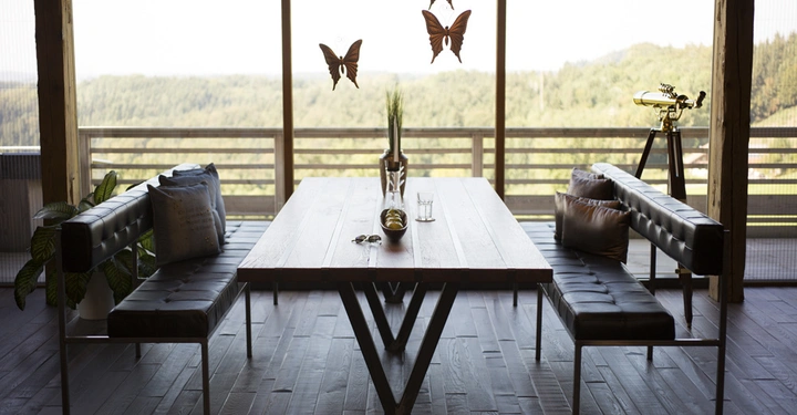 Galerie-Bild 3: Holztisch steht auf einem Holzfußboden mit zwei Ledersofas davor. Glasvase auf dem Tisch. Hintergrund aus großen Fenstern mit Blick auf dichte grüne Wälder. Hängende Schmetterlingsdekorationen.