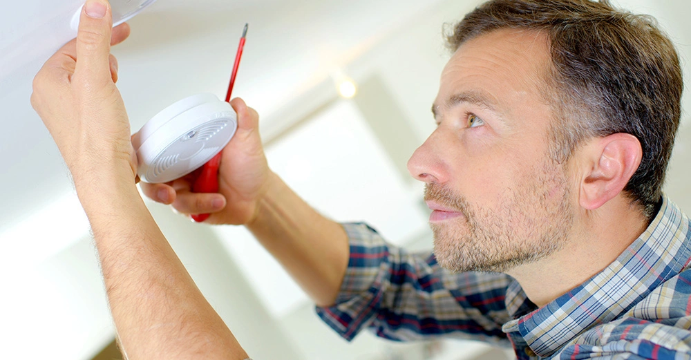 Mann installiert Rauchmelder an der Decke mit Schraubenzieher in der Hand im hellen Raum mit Fenstern im Hintergrund.