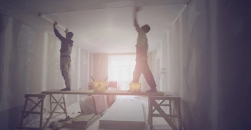 Zwei Bauarbeiter arbeiten an der Decke eines Raumes auf einem erhöhten Gerüst in einem im Bau befindlichen Gebäude mit spärlicher Beleuchtung und Wänden in unterschiedlichen Bauzuständen.