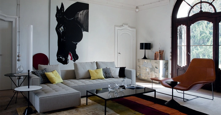 Graues Sofa mit Kissen steht in einem stilvollen Wohnzimmer vor einem großformatigen schwarzen Pferdebild Hölzerne Kommode und schwarzer Sessel in der Nähe von großen Fenstern