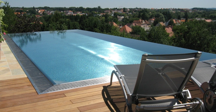 Ein Liegestuhl steht still auf einem Holzdeck neben einem Infinity-Pool, der über den Rand hinauszugehen scheint; im Hintergrund eine hügelige Landschaft und ein Dorf mit Häusern und Bäumen.