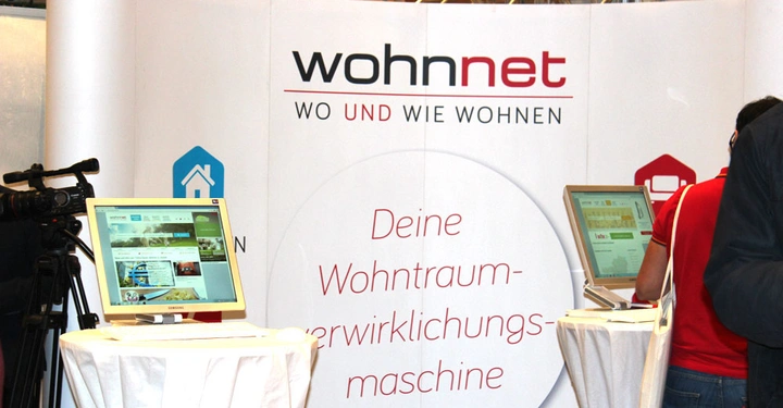 Galerie-Bild 11: Zwei Computerbildschirme zeigen Webseiten an, Menschen bedienen die Bildschirme im Kontext einer Wohnmesse, umgeben von einem Wohnnet-Stand. Text: Wohnnet WO UND WIE WOHNEN Deine Wohntraumverwirklichungsmaschine.