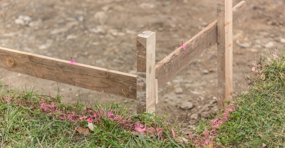 Holzpfosten stehen im Boden und sind mit pinkem Band markiert, umgeben von Gras und Erde in einer Baustellenumgebung.