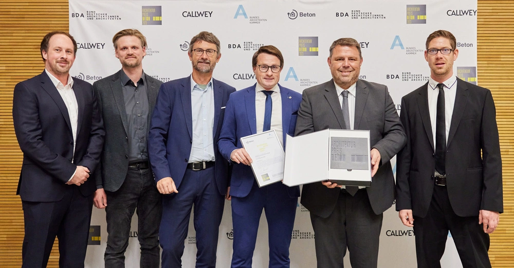 Sechs Männer tragen Anzüge und lächeln in die Kamera. Zwei halten Zertifikate und einen Preis. Sie stehen vor einer Wand mit Logos in einem gut beleuchteten Raum.