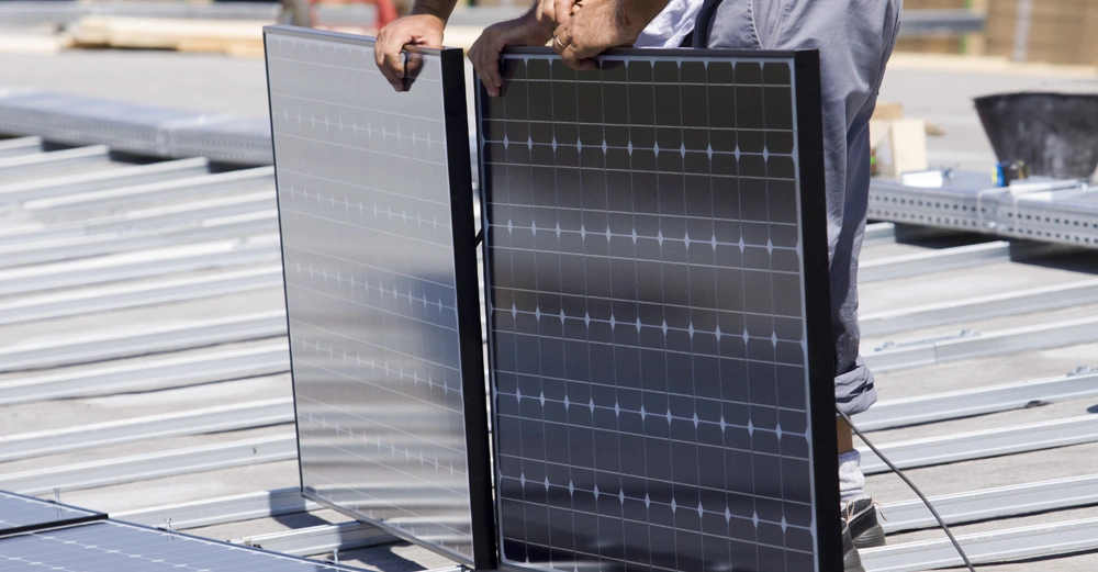 Eine Person installiert zwei Solarpaneele auf einem Dach in einem industriellen Umfeld begleitet von anderen Baumaterialien im Hintergrund