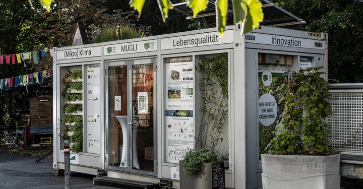 Containergebäude mit Pflanzen an den Wänden und Türen auf einem Parkplatz umgeben von Bäumen und einer farbenfrohen Wimpelkette. Text auf dem Gebäude Mikro Klima Lebensqualität Innovation MUGLI. Schild Hier entsteht eine neue grüne Wand.