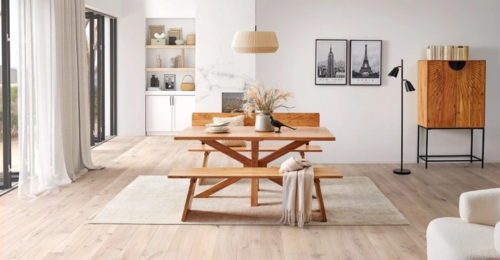 Moderner, heller Essbereich mit Massivholztisch und Holzbänken auf einem beigen Teppich. Im Hintergrund ein Holzschrank mit Sonnenstrahl-Muster, Schwarz-Weiß-Bilder und eine schwarze Stehlampe. Lichtdurchfluteter Raum mit hellem Echtholzboden.