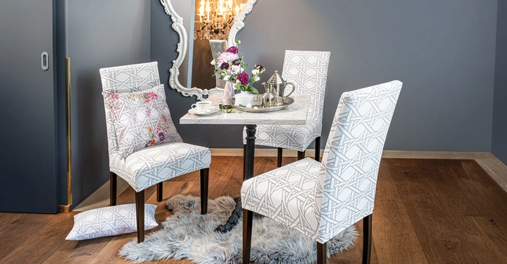 Vier gepolsterte Stühle mit geometrischem Muster stehen um einen Tisch mit Blumen und Teeservice vor einem großen Spiegel in einem modern eingerichteten Raum mit Holzboden und grauen Wänden.