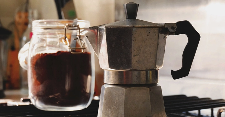 Ein silbernes Mokkakännchen steht auf einem Herd neben einem großen Glasbehälter mit Kaffeepulver in einer häuslichen Küchenszene.