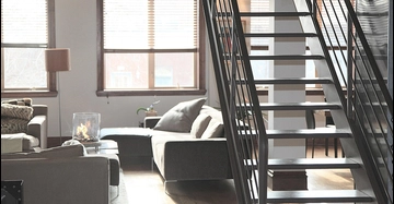 Helles Loft-Wohnzimmer mit grauem Sofa, Couchtisch mit Glasfeuer, großen Fenstern und schwarzer Metalltreppe im Industrial-Stil.