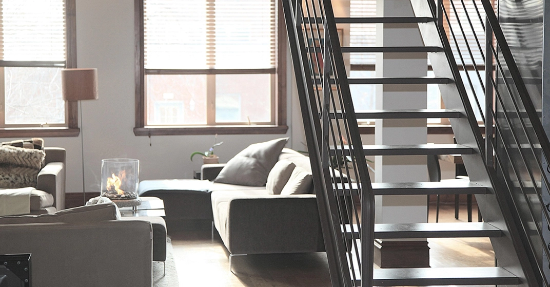 Helles Loft-Wohnzimmer mit grauem Sofa, Couchtisch mit Glasfeuer, großen Fenstern und schwarzer Metalltreppe im Industrial-Stil.