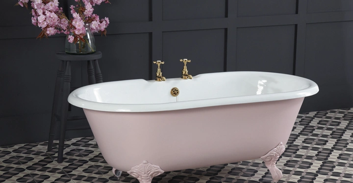 Rosa Badewanne mit weißen Innenwänden und goldenen Armaturen steht auf dekorativen Füßen auf schwarz-weiß gemusterten Fliesen in einem dunklen Raum Ein Hocker mit einer Vase voll rosa Blüten steht daneben