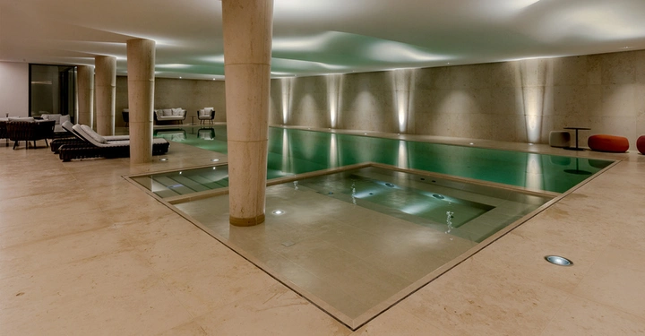 Schwimmbecken mit angrenzendem Whirlpool steht still im Raum mit beige gefliestem Boden und mehreren Sitzgelegenheiten sowie Säulen die die Decke stützen in einem modernen Innenbereich.