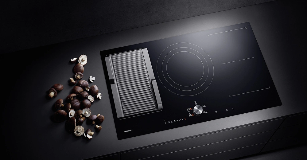 Ein modernes Kochfeld mit einem runden Brenner links und rechts sowie einem Grill in der Mitte liegt auf einer dunklen Küchenarbeitsplatte. Links daneben liegen geschnittene Kastanien.
