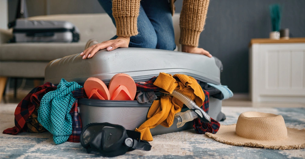 Frau versucht extrem vollen Reisekoffer zu schließen. Koffer liegt auf grauem Teppichboden, Utensilien wie Taucherbrille, Sonnenhut und Shirts lugen hervor