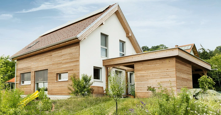 Ein zweistöckiges Holzhaus mit Solarpanelen auf dem Dach befindet sich in einer grünen Landschaft umgeben von Bäumen und Wiese.
