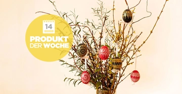 Glasvase mit verzierten Ostereiern hängt an Zweigen in einem hell beleuchteten Raum Tisch. Text: Produkt der Woche 14