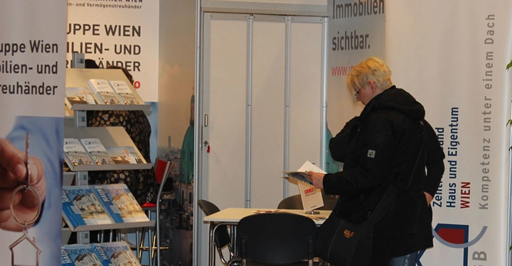 Galerie-Bild 15: Eine Person liest ein Dokument in einem Messestand mit Broschürenregalen und Plakaten der Wirtschaftskammer Wien für Immobilien- und Vermögensverwalter. Text Wir machen Ihren Immobilien sichtbar.