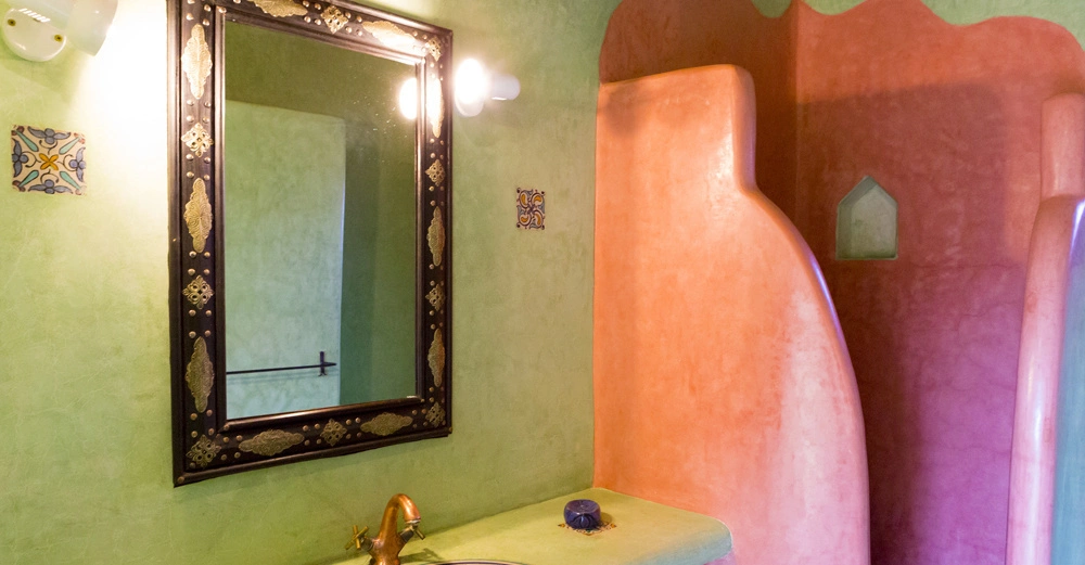 Spiegel hängt an einer grünlichen Wand und reflektiert den Raum Handwaschbecken mit goldfarbenem Wasserhahn darunter neben einer rosa Kurve in einem rustikal dekorierten Badezimmer.