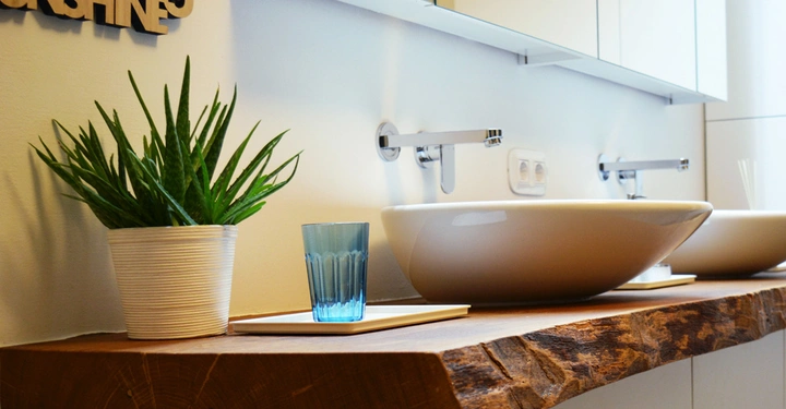 Topfpflanze steht neben einem Glas auf einem rustikalen Waschtisch mit zwei runden Waschbecken in einem modernen Badezimmer. Wanddekoration aus Holz mit der Aufschrift SUNSHINE9 an der Wand.
