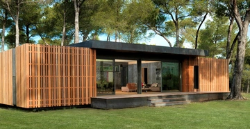 Modernes einstöckiges Haus mit Holzverkleidung geöffneten Glasschiebetüren und Terrasse umgeben von Rasen und hohen Bäumen im Freien.