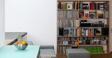 Eine Metallschüssel mit Früchten steht auf einem türkisfarbenen Tisch in einem Raum mit einem grauen Sofa und einem großen Bücher- und Plattenregal an einer weißen Wand