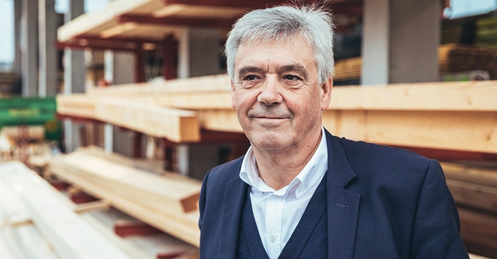 Ein Mann steht lächelnd in Geschäftskleidung in einem Holzlager mit gestapeltem Bauholz im Hintergrund.