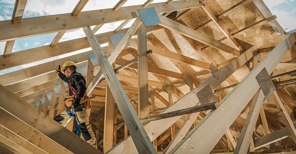 Bauarbeiter trägt Helm und Werkzeuggürtel arbeitet an Holzrahmenkonstruktion umgeben von Holzbalken und Dachlatten unter freiem Himmel.