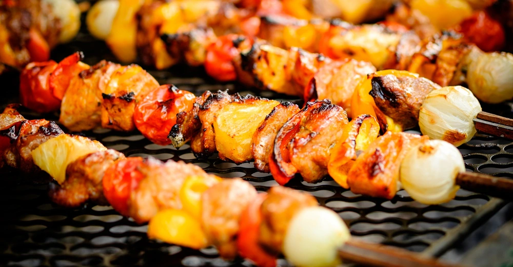 Mehrere Spieße mit gegrillten Fleischstücken, Paprika, Zwiebeln und Ananas auf einem Grillrost, umgeben von weiteren gegrillten Lebensmitteln in verschwommenem Hintergrund.