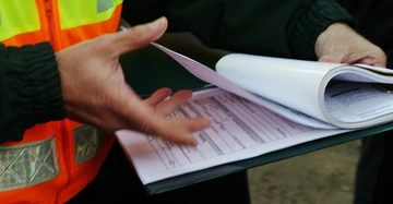 Bauarbeiter mit knallgelb und roter Warnweste, man sieht nur seinen Oberkörper und die Hände, hält ein Klemmbrett mit Formularen, die er durchblättert.