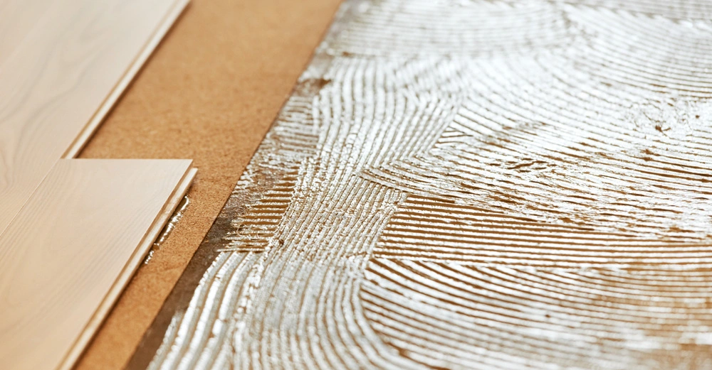 Holzbodenplatten werden auf eine Klebefläche mit gezackter Struktur gelegt. Links sind die Platten. Rechts ist der Kleber. Sie befinden sich auf einem Holzuntergrund.