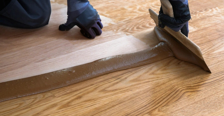 Eine Person mit Handschuhen verteilt braunes Holzfinish mit einem Spachtel auf einem Holzboden in einem Innenraum.