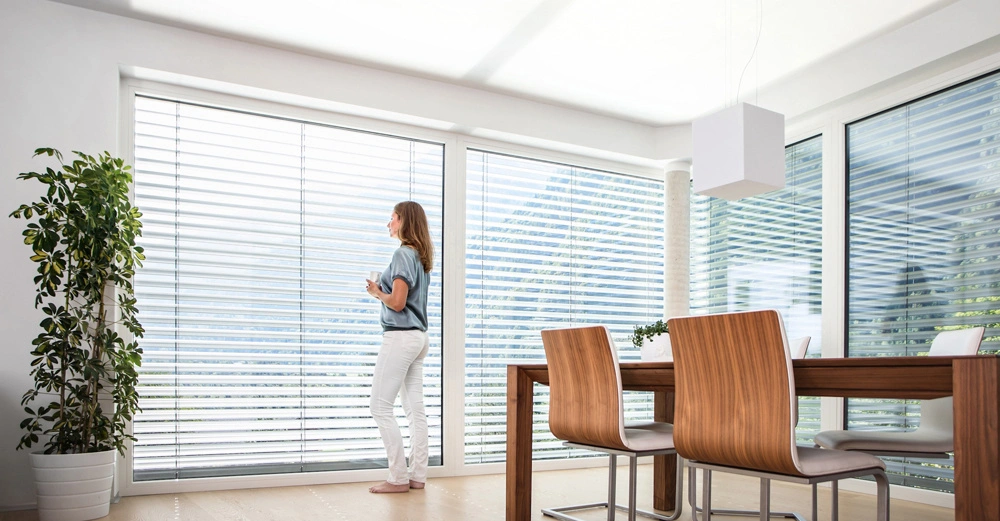 Frau steht vor großen Fenstern mit Jalousien hält eine Tasse und schaut hinaus modernes Esszimmer mit Holztisch vier Stühlen und Topfpflanze helle und minimalistische Einrichtung.