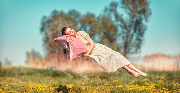 Frau schläft auf einem rosa Kissen in der Luft schwebend auf einer blumigen Wiese mit Bäumen im Hintergrund.
