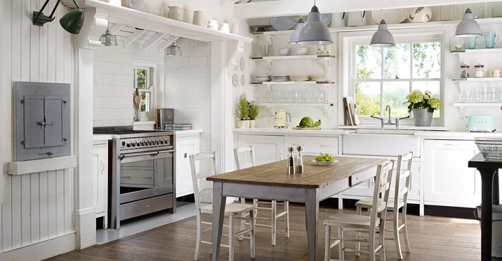 Galerie-Bild 1: Ein rustikaler Küchentisch mit vier Stühlen steht im Zentrum einer hellen Küche mit weißen Schränken, Regalen voller Geschirr, und einem großen Fenster mit Blick ins Grüne. Links ein Herd.