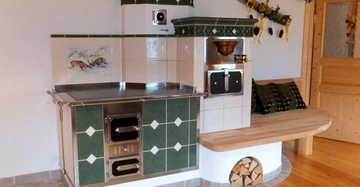 Ein grüner Kachelofen mit weißen Details ist in einer Ecke eines Raumes platziert. Auf dem Ofen ist ein Gemälde von zwei Rehen in einer Berglandschaft. Daneben eine Holzbank mit Kissen.