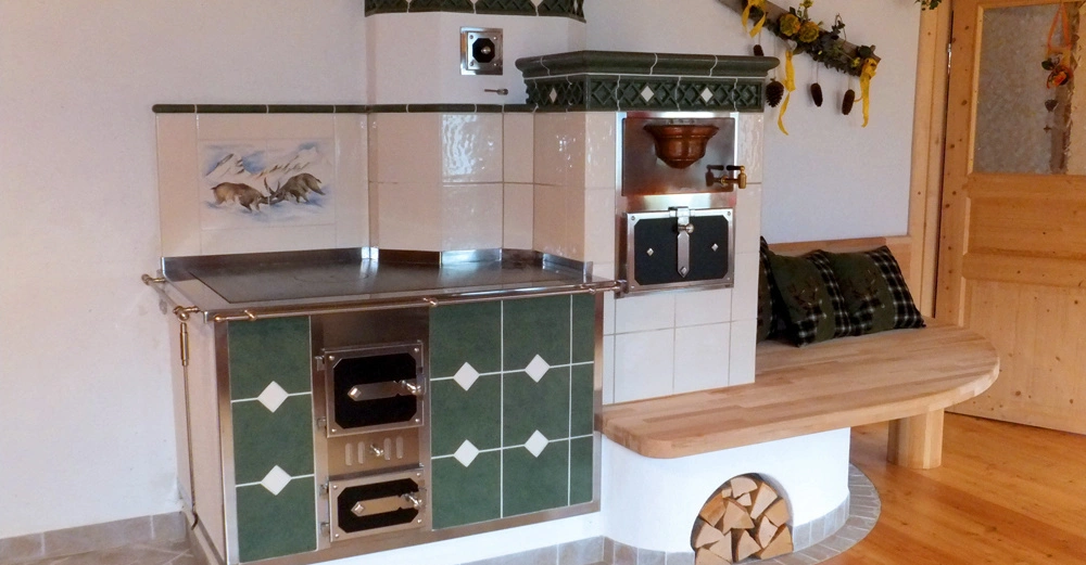 Ein grüner Kachelofen mit weißen Details ist in einer Ecke eines Raumes platziert. Auf dem Ofen ist ein Gemälde von zwei Rehen in einer Berglandschaft. Daneben eine Holzbank mit Kissen.