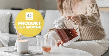 Eine Person gießt Tee aus einem gläsernen Krug in Gläser auf einem Tisch in einem gemütlichen Wohnzimmer. Text auf gelbem Kreis: 42 Produkt der Woche.
