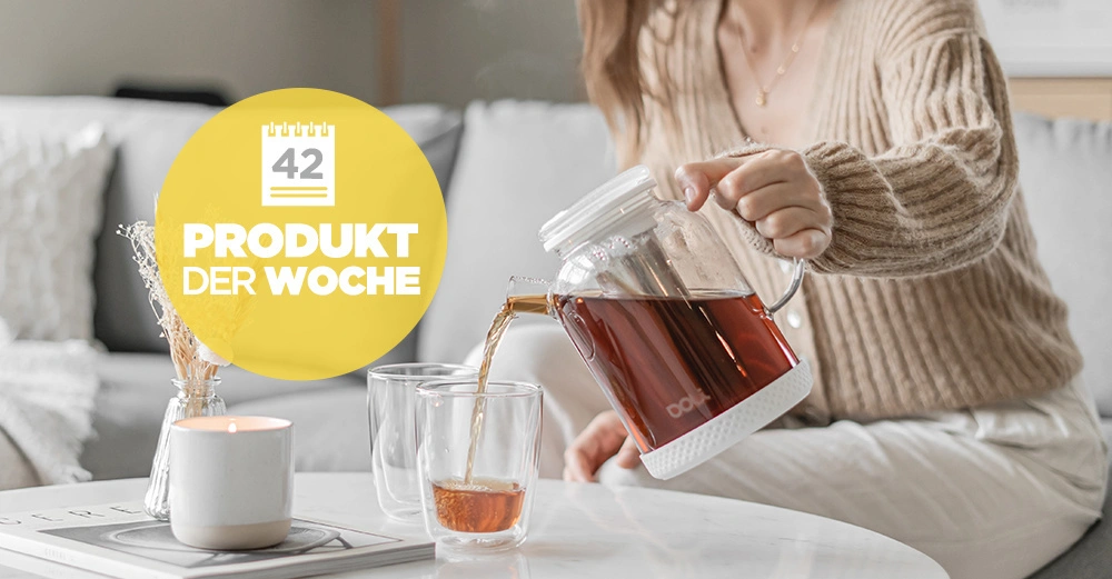 Eine Person gießt Tee aus einem gläsernen Krug in Gläser auf einem Tisch in einem gemütlichen Wohnzimmer. Text auf gelbem Kreis: 42 Produkt der Woche.