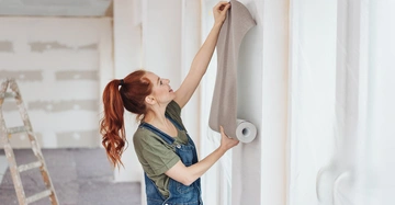 Fröhliche rothaarige Frau in einer blauen Jeanslatzhose installiert lächelnd eine graue Tapete an einer weißen Wand.
