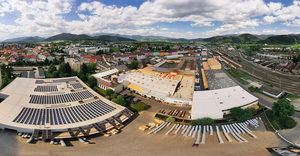 Fabrikgebäude mit Solarpaneelen auf dem Dach steht in einer Kleinstadt umgeben von Wohnhäusern und Bahngleisen vor einer bergigen Landschaft bei bewölktem Himmel