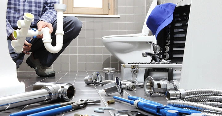 Galerie-Bild 2: Eine Person installiert Rohrleitungen kniend in einem Badezimmer umgeben von Werkzeugen und Sanitärteilen mit einem Werkzeugkasten neben einer Toilette.